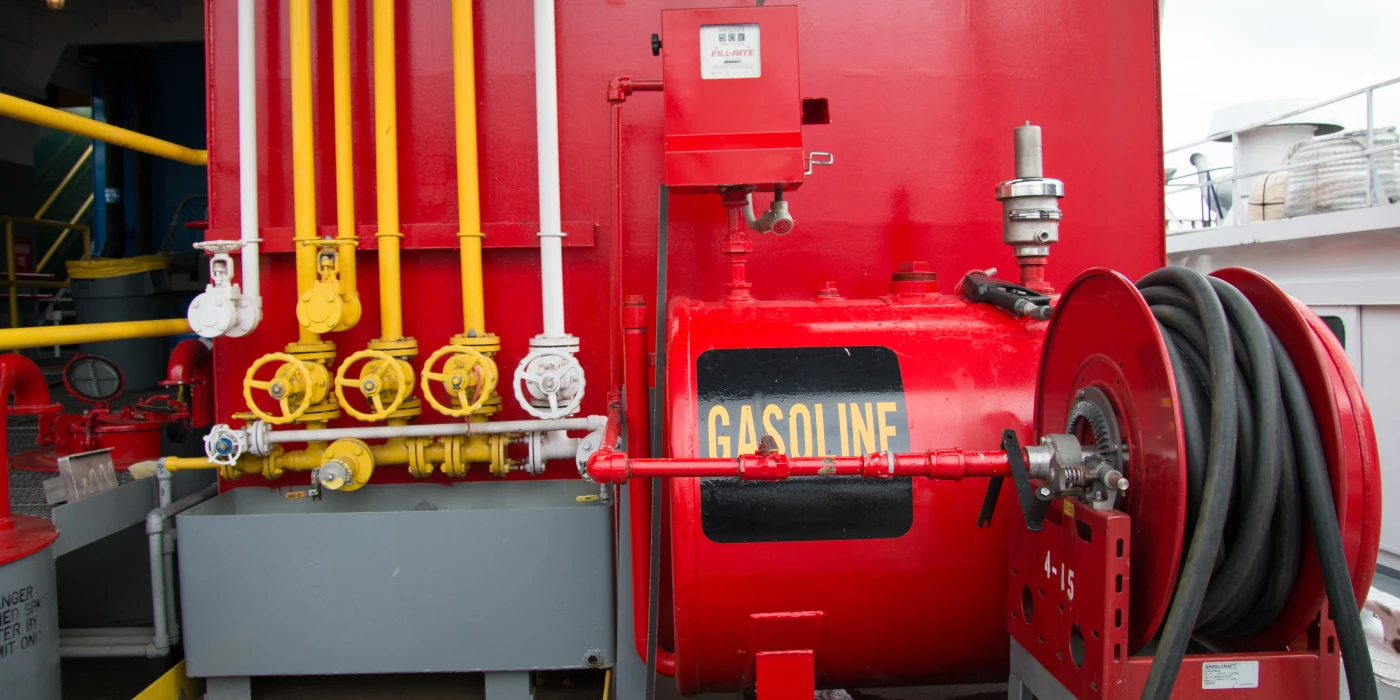 Red gasoline storage tank with labeled pipes, valves, and a hose reel on an industrial platform.
