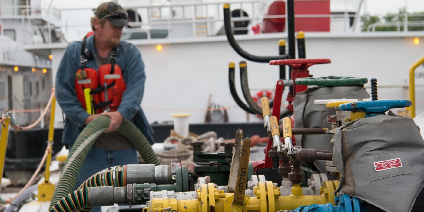 A worker in a life vest handles hoses connected to industrial valves and machinery on a docked vessel.