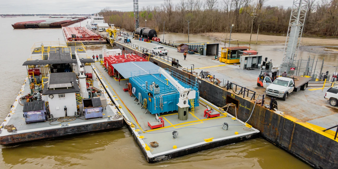Dock workers oversee loading of large blue industrial equipment onto a barge at a riverside shipping facility.