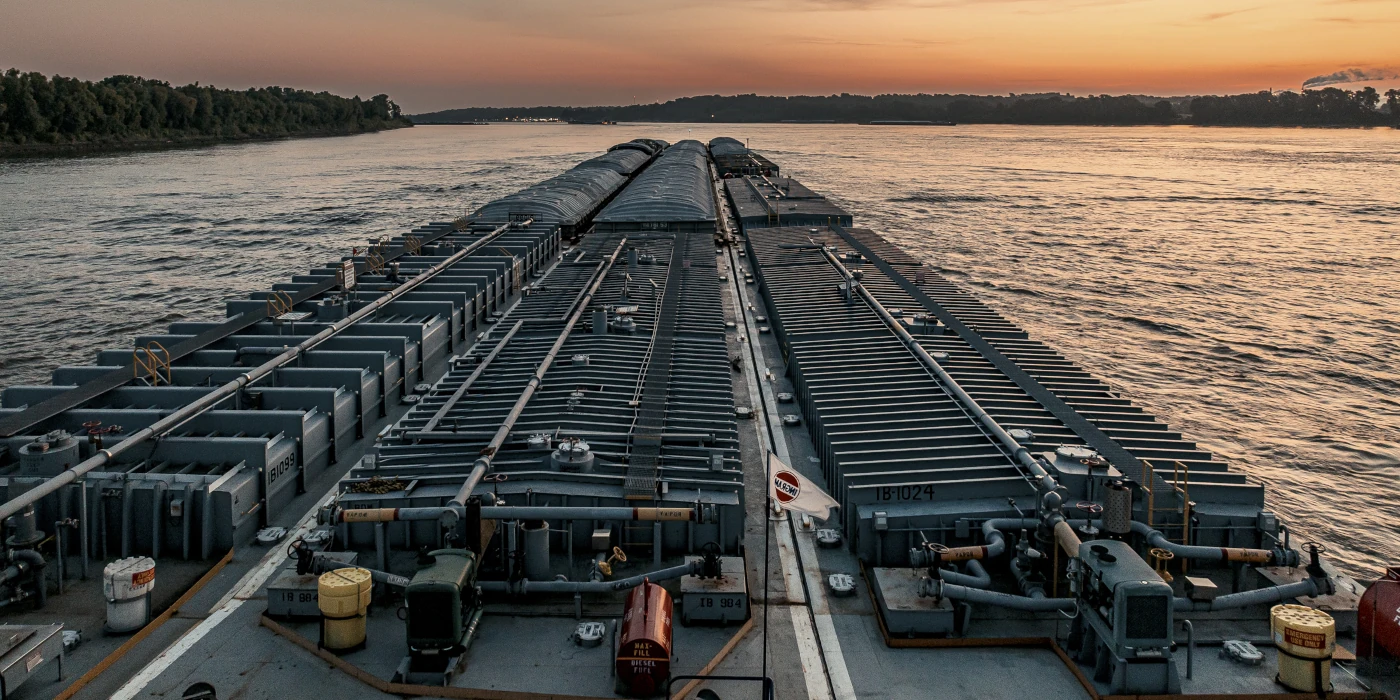 A barge carrying cargo travels along a wide river at sunset, with trees lining the distant shoreline.