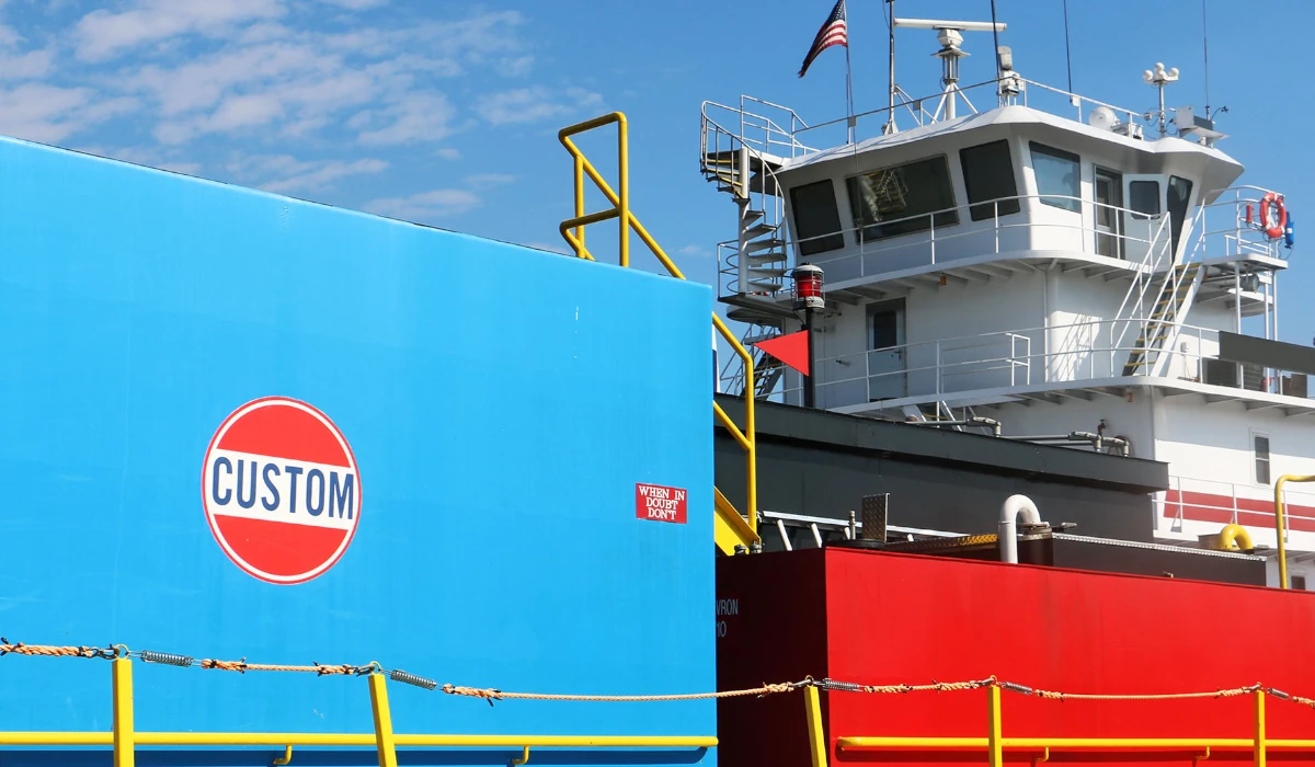 A tugboat with blue and red containers labeled CUSTOM docked under a clear, partly cloudy sky.