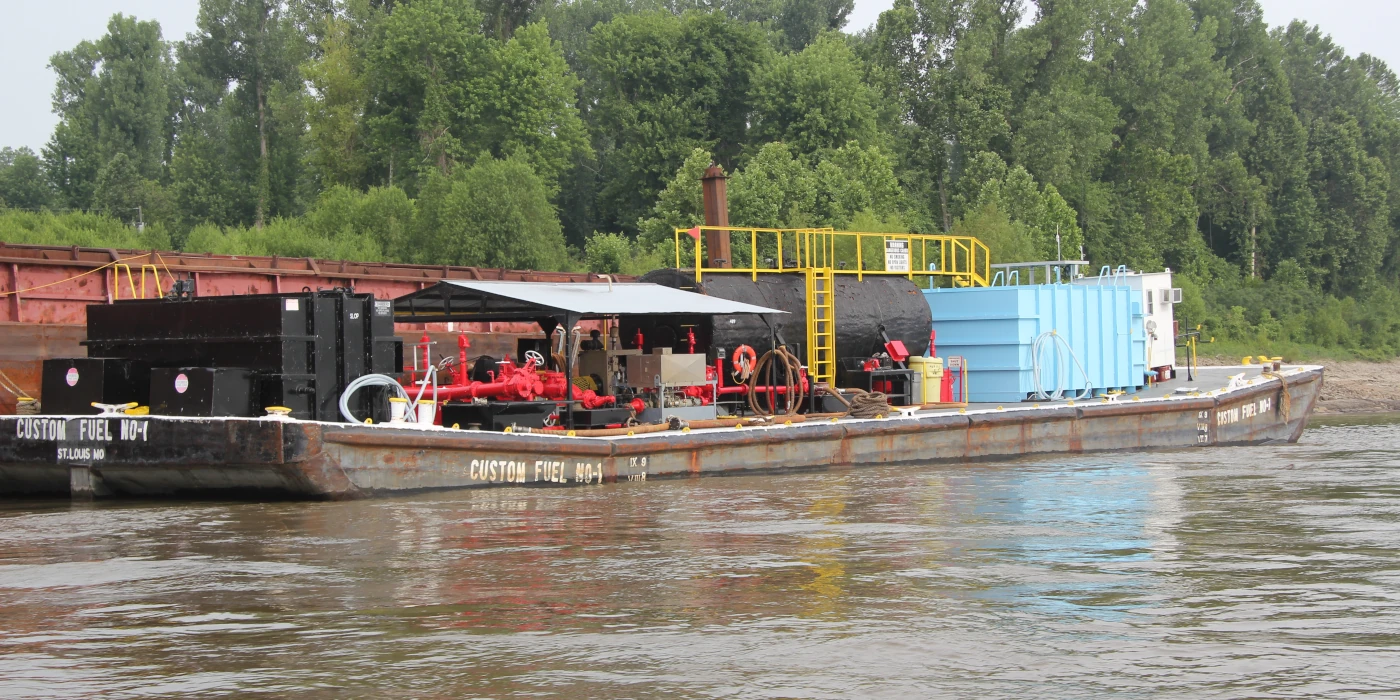 A flat barge carrying industrial fuel equipment and machinery is docked near a forested riverbank.