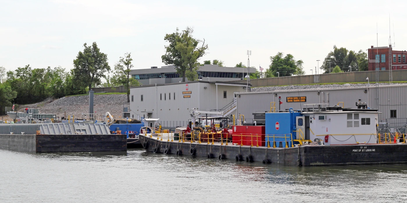 Several industrial barges are docked alongside a waterfront facility with equipment and buildings nearby.