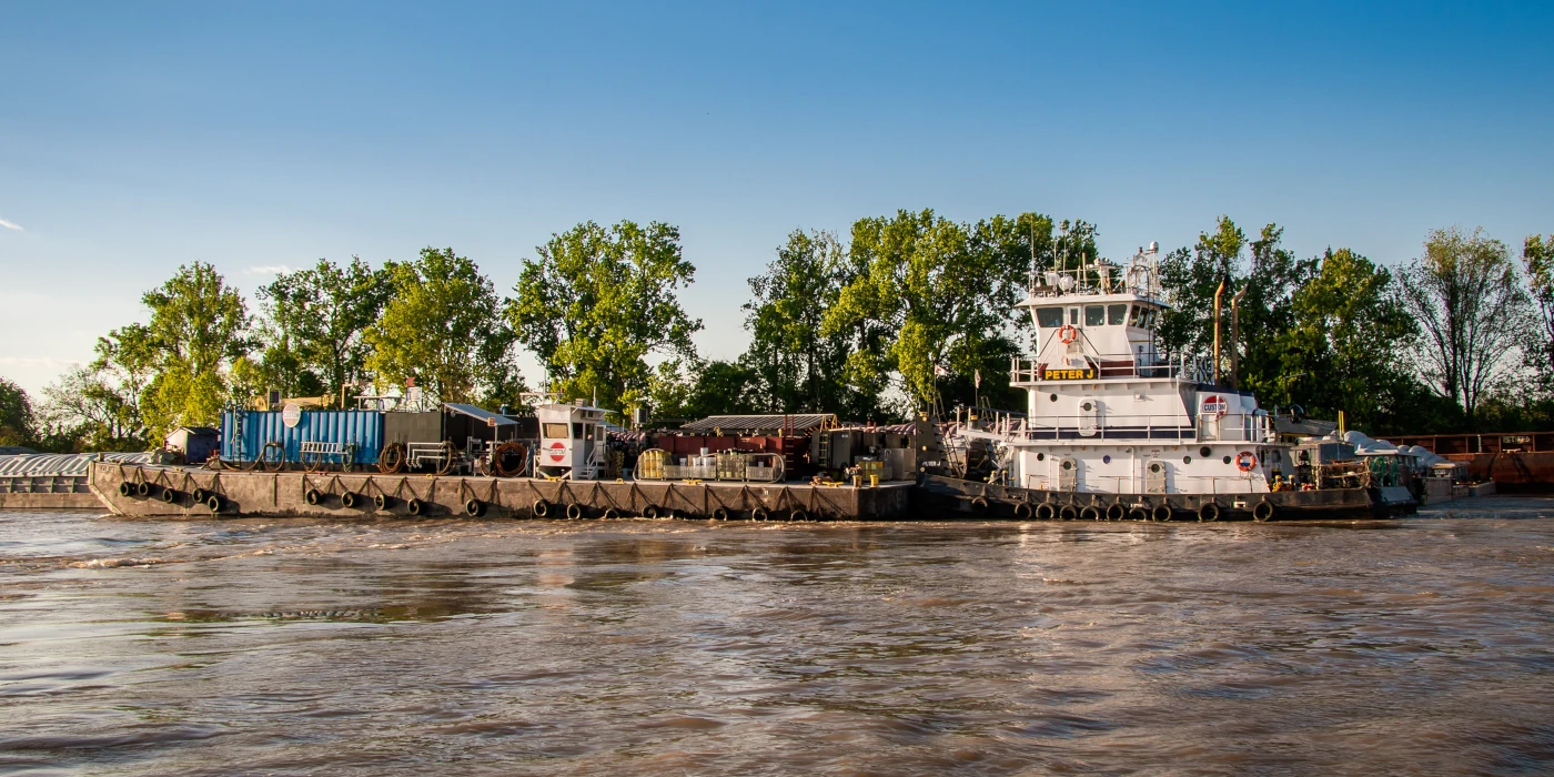 A tugboat pulls a loaded barge along a river with trees visible in the background under a clear sky.